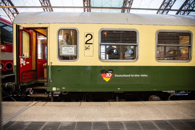 A yellow and green historical second-class railway carriage sits on the tracks in a train station. The railway carriage displays the logo of the anniversary year with the words „Deutschland ist eins: vieles” (Germany: coming together as one)