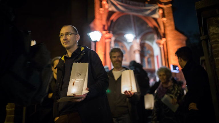 In front of the illuminated entrance of the Gethsemanekirche church in Berlin, people hold paper lanterns in one hand.