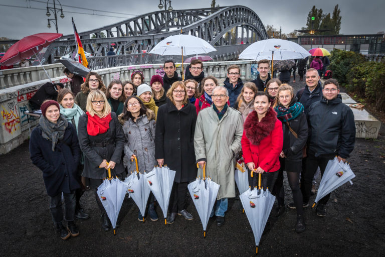 A group of people stand in front of a bridge, the Bösebrücke, at the former border crossing point at Bornholmer Straße in Berlin. The weather is rainy and some of them hold umbrellas with the logo of the anniversary year.