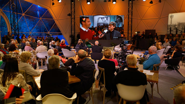 In a television studio, people sit at tables looking at a large screen. On the screen, a man is being interviewed.