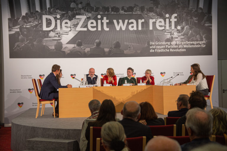 Eight people sit on a stage in front of a backdrop bearing the words „Die Zeit war reif”, meaning „The time had come”. Microphones are positioned in front of them.