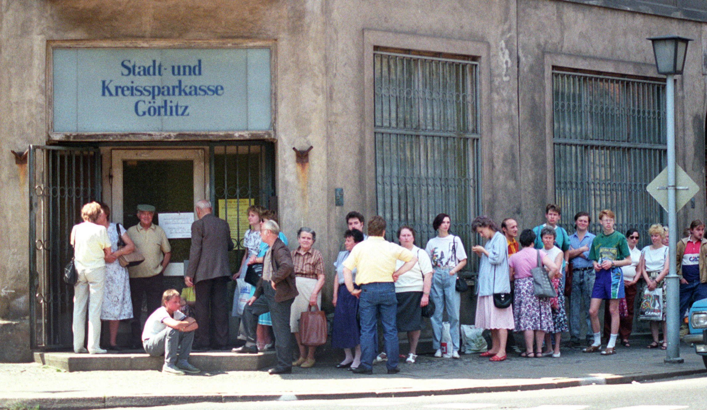 Eine lange Schlange von Menschen wartet vor einem Eckgebäude. Über dem Eingang steht ein Schild mit der Aufschrift "Stadt- und Kreissparkasse Görlitz".